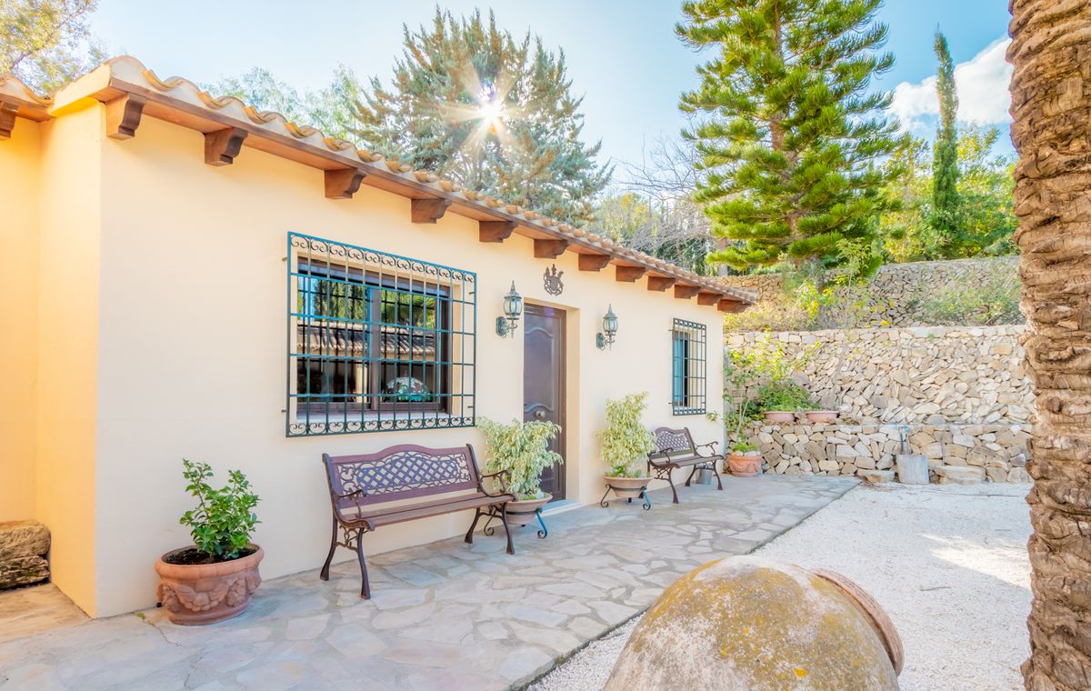 Fachada mediterránea con vigas de madera expuestas, ventanas con rejas de hierro forjado y un patio de piedra natural con bancos ornamentales.