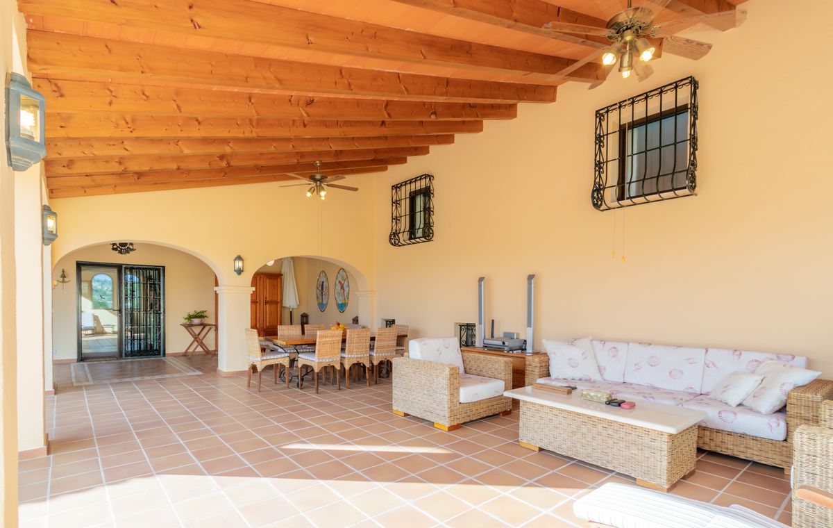 Terraza cubierta con vigas de madera a la vista, suelos de terracota y mobiliario de mimbre que incluye zona de estar y comedor exterior.