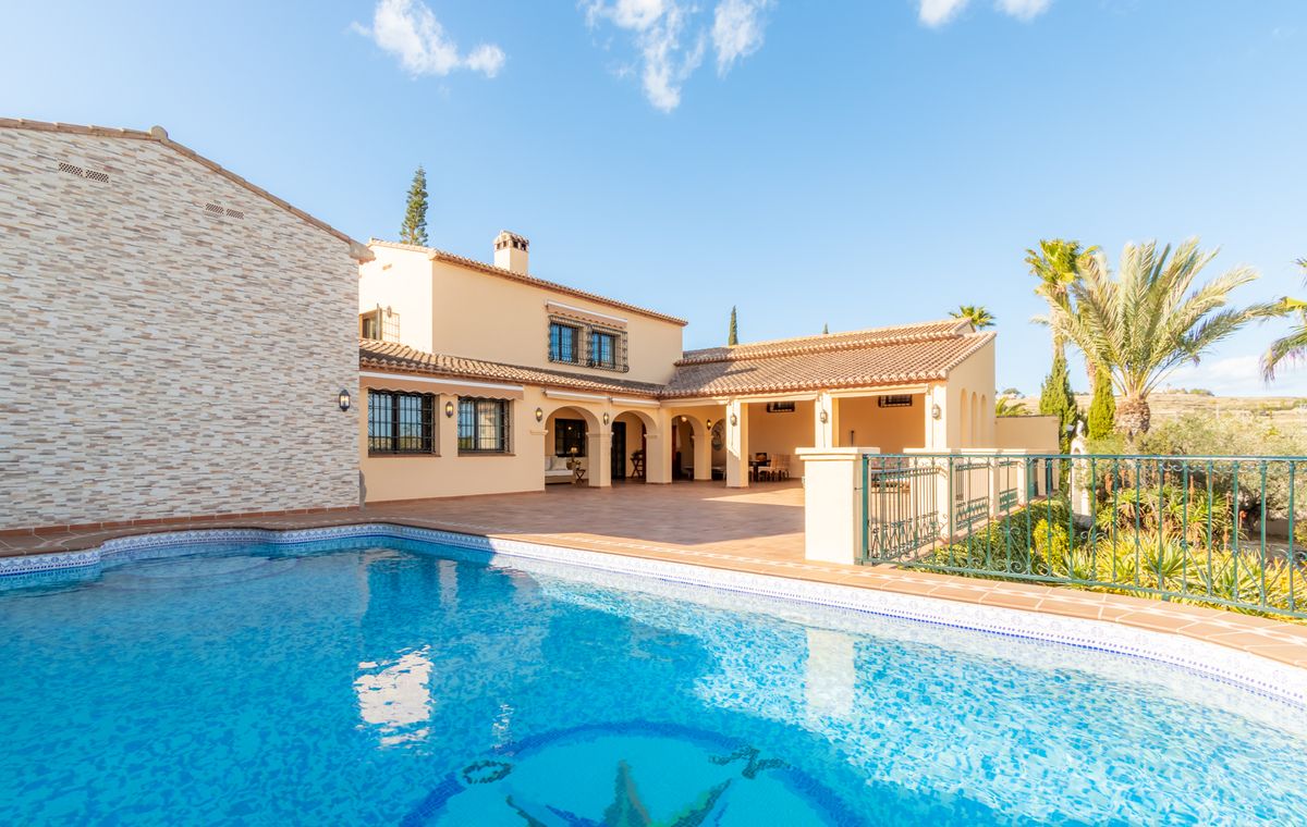 Vista exterior de una villa mediterránea con piscina de mosaico, amplia terraza de terracota y porche cubierto con arcos tradicionales.