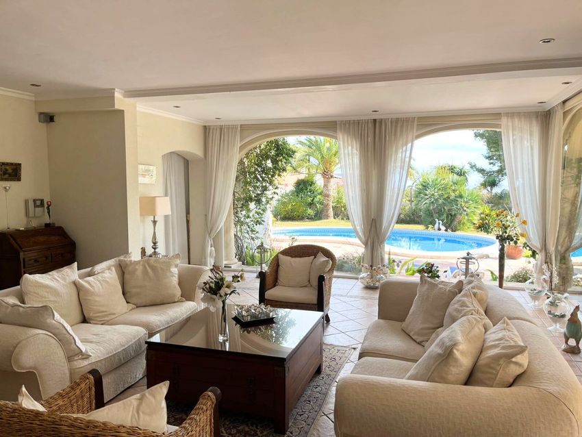 Amplio salón con grandes ventanales en arco que ofrecen vistas directas a la piscina y al exuberante jardín mediterráneo. Sofás claros y mesa de centro de madera con cristal complementan el ambiente luminoso. Amplio salón con grandes ventanales en arco que ofrecen vistas directas a la piscina y al exuberante jardín mediterráneo. Sofás claros y mesa de centro de madera con cristal complementan el ambiente luminoso.