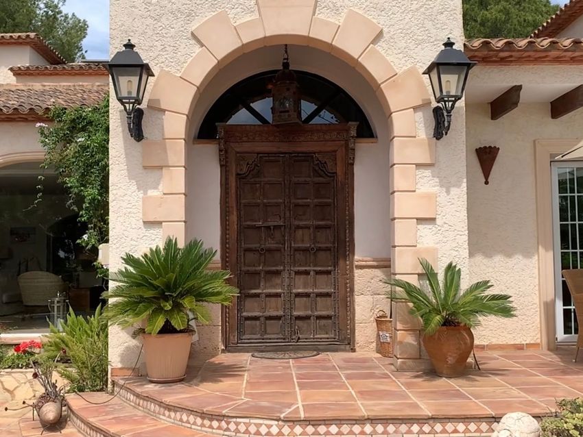 Entrada principal con una gran puerta doble de madera tallada bajo un arco de piedra, flanqueada por faroles y plantas en maceta sobre escalones de terracota. Entrada principal con una gran puerta doble de madera tallada bajo un arco de piedra, flanqueada por faroles y plantas en maceta sobre escalones de terracota.