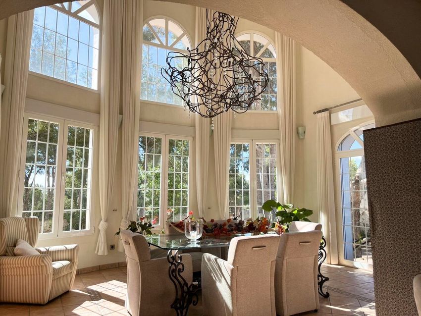 Este comedor presenta un techo de doble altura y grandes ventanales en arco con celosía, que inundan el espacio de luz natural. Una distintiva lámpara de diseño cuelga sobre una mesa de cristal con base de metal forjado. Este comedor presenta un techo de doble altura y grandes ventanales en arco con celosía, que inundan el espacio de luz natural. Una distintiva lámpara de diseño cuelga sobre una mesa de cristal con base de metal forjado.
