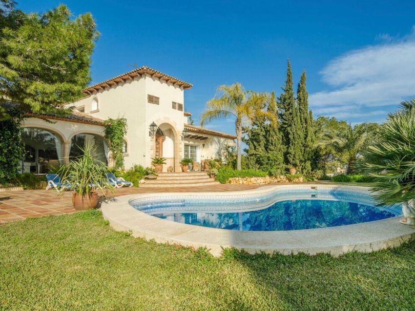 Exterior de villa mediterránea con piscina de forma irregular y terraza de baldosas. El jardín presenta césped verde y árboles maduros, bajo un cielo azul despejado. Exterior de villa mediterránea con piscina de forma irregular y terraza de baldosas. El jardín presenta césped verde y árboles maduros, bajo un cielo azul despejado.