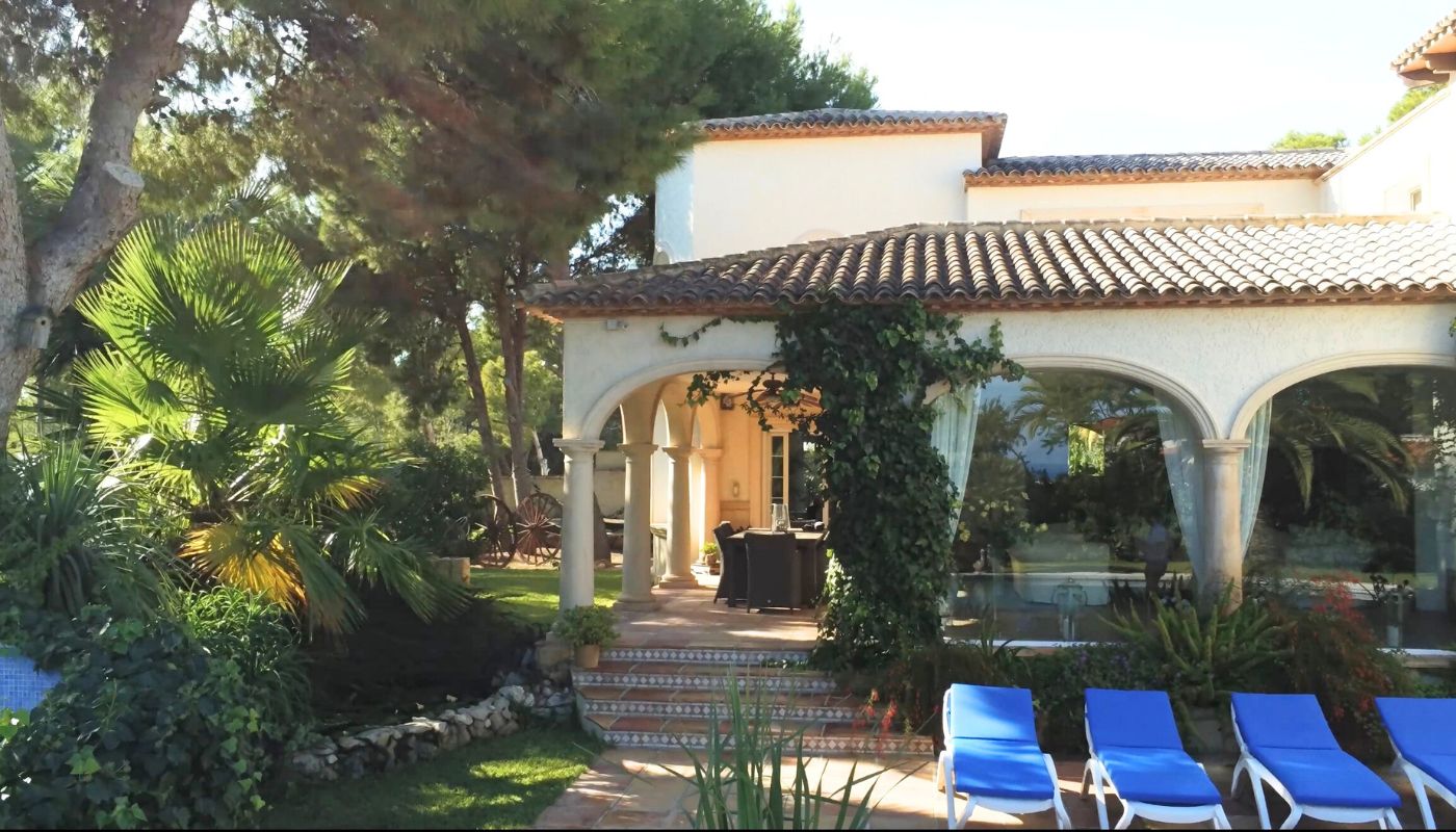 Exterior de villa mediterránea con porche de arcos y columnas de piedra, tejado de teja árabe y terraza con tumbonas azules junto al jardín.