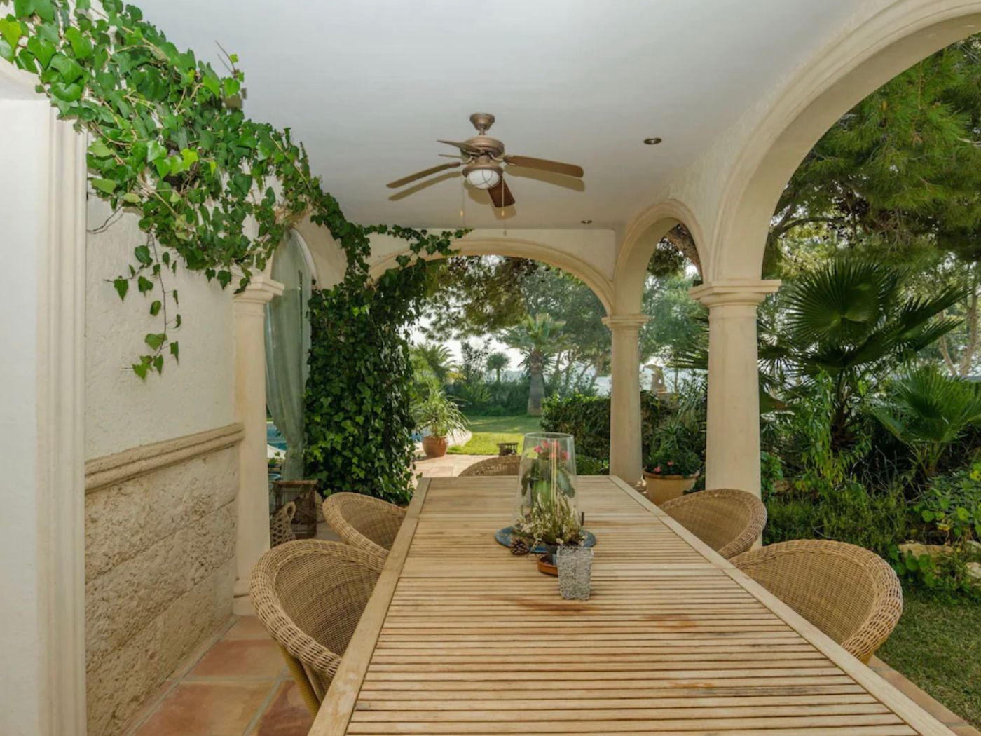 Porche cubierto con arcos de piedra tosca, gran mesa de madera y sillas de mimbre, integrado en un jardín con hiedra y vegetación mediterránea.