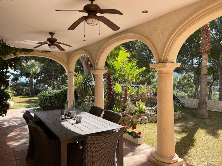 Terraza cubierta de estilo mediterráneo con arcos y columnas, equipada con un comedor exterior de mimbre y ventiladores de techo. Ofrece vistas al exuberante jardín y un atisbo del mar. Terraza cubierta de estilo mediterráneo con arcos y columnas, equipada con un comedor exterior de mimbre y ventiladores de techo. Ofrece vistas al exuberante jardín y un atisbo del mar.