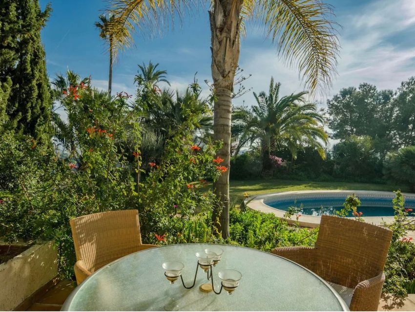 Zona de comedor exterior con mesa de cristal y sillas de mimbre, rodeada de exuberante vegetación mediterránea y una piscina curva al fondo. Zona de comedor exterior con mesa de cristal y sillas de mimbre, rodeada de exuberante vegetación mediterránea y una piscina curva al fondo.