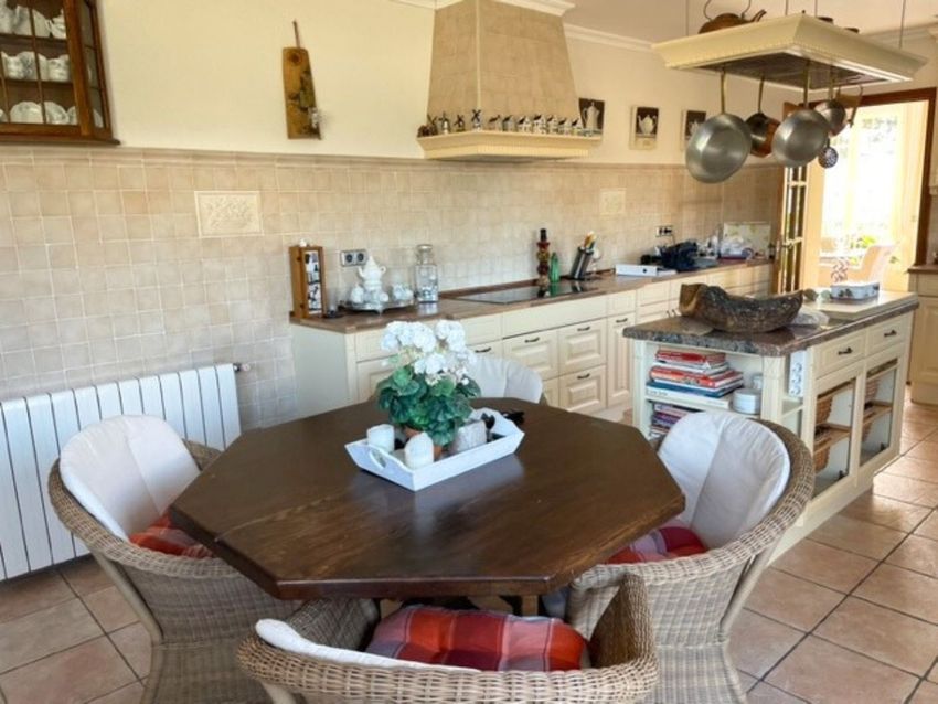 Zona de desayuno con mesa hexagonal de madera y sillas de mimbre, junto a una cocina rústica equipada con isla central y encimera de granito. Zona de desayuno con mesa hexagonal de madera y sillas de mimbre, junto a una cocina rústica equipada con isla central y encimera de granito.