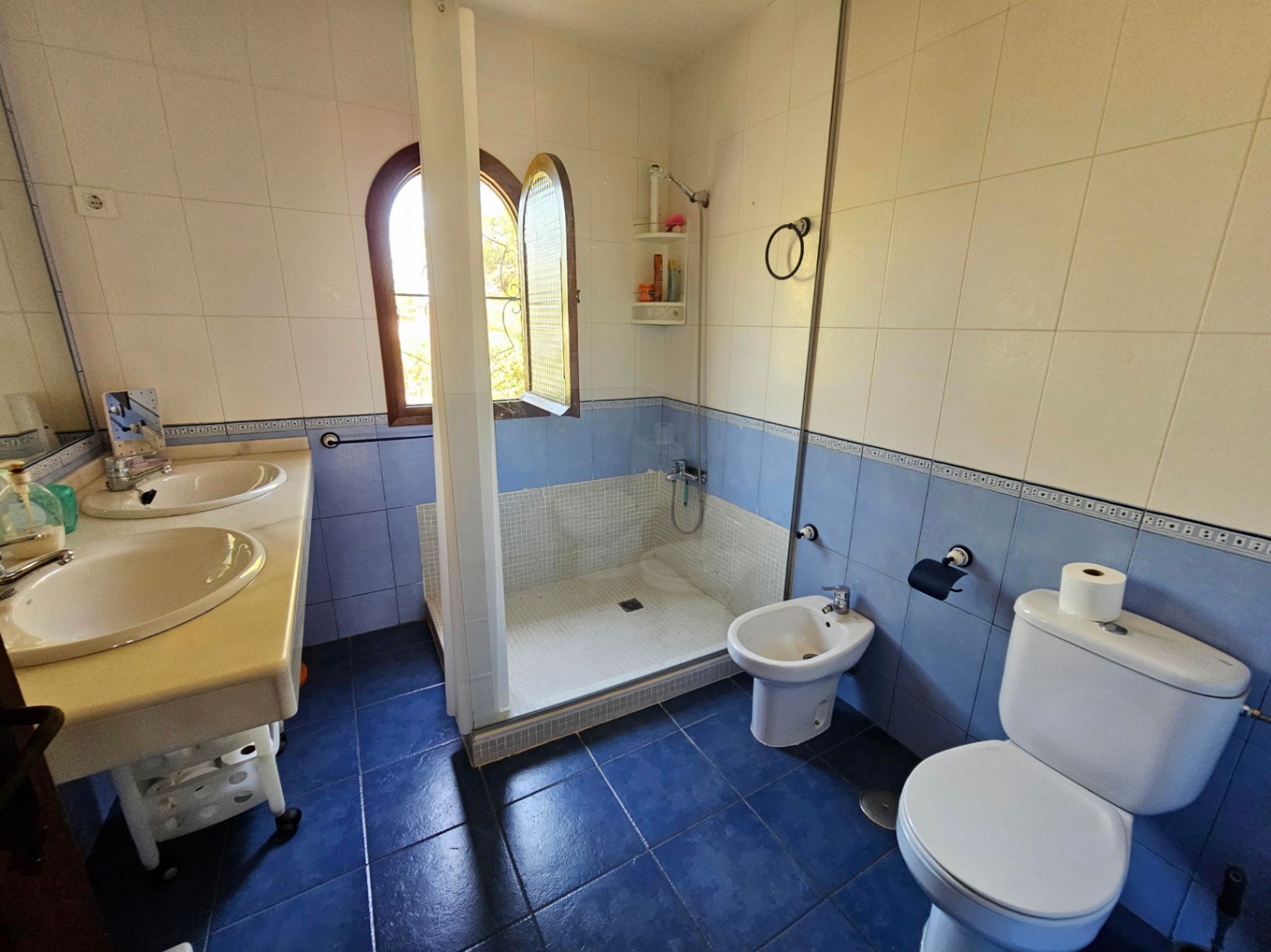 Baño con lavabo doble de mármol, ducha de obra con mosaico, bidé e inodoro. Destaca la ventana de arco de madera y el alicatado bitono en azul y blanco.