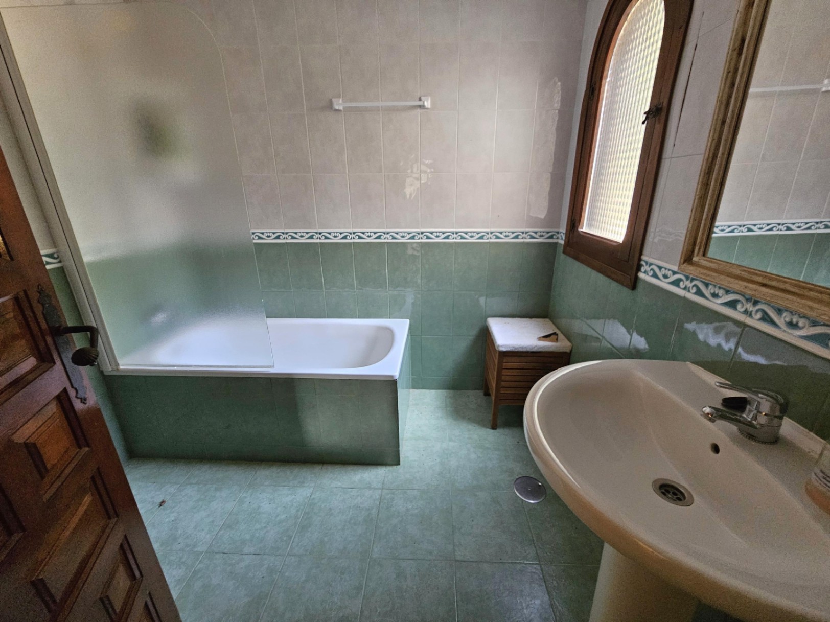 Baño de estilo tradicional con azulejos verdes y blancos, bañera integrada con mampara de cristal esmerilado y ventana de madera con arco de medio punto.