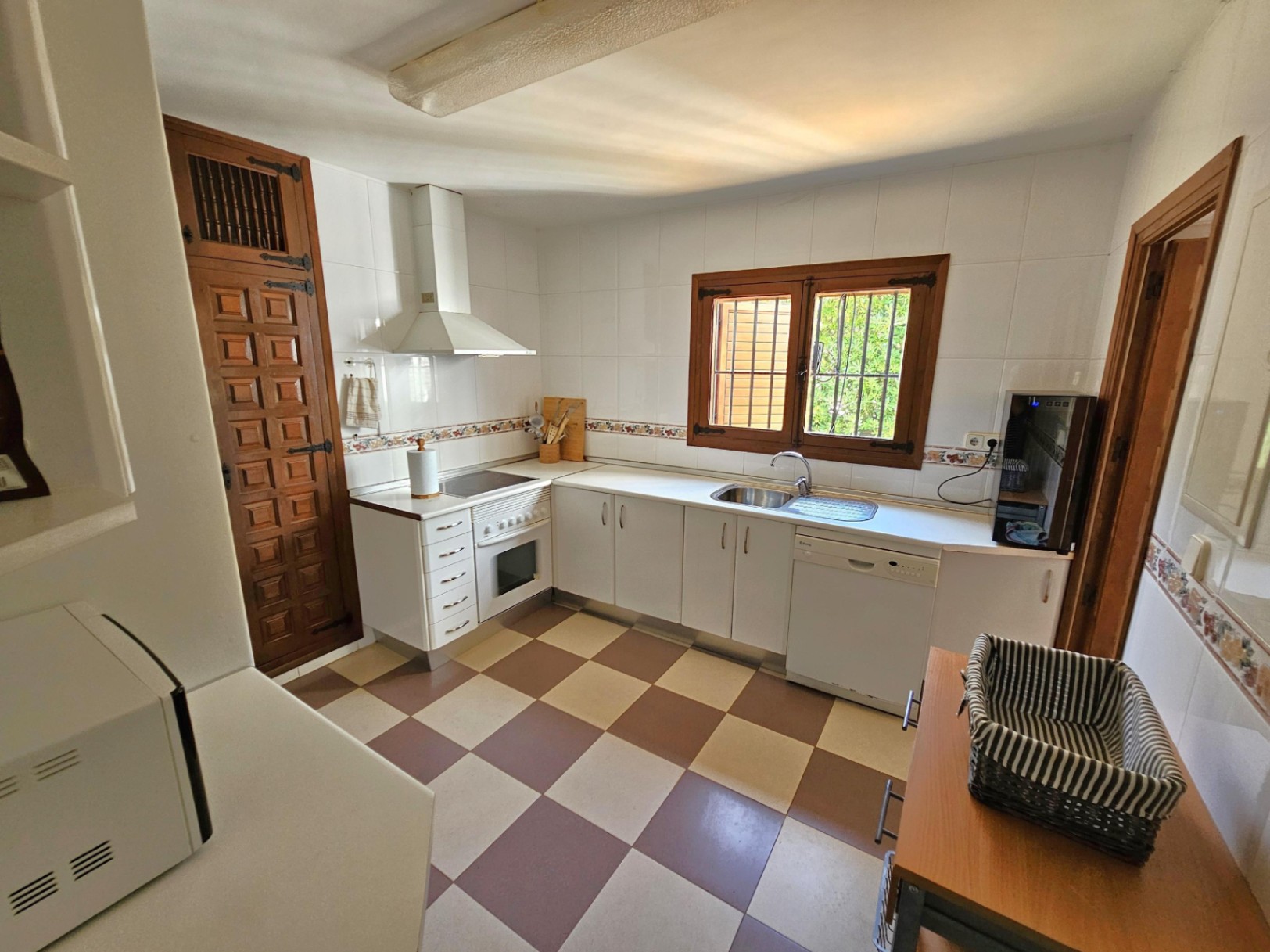 Cocina con armarios blancos, suelo damero en tonos tierra y carpintería de madera oscura. Dispone de electrodomésticos integrados y encimera blanca.