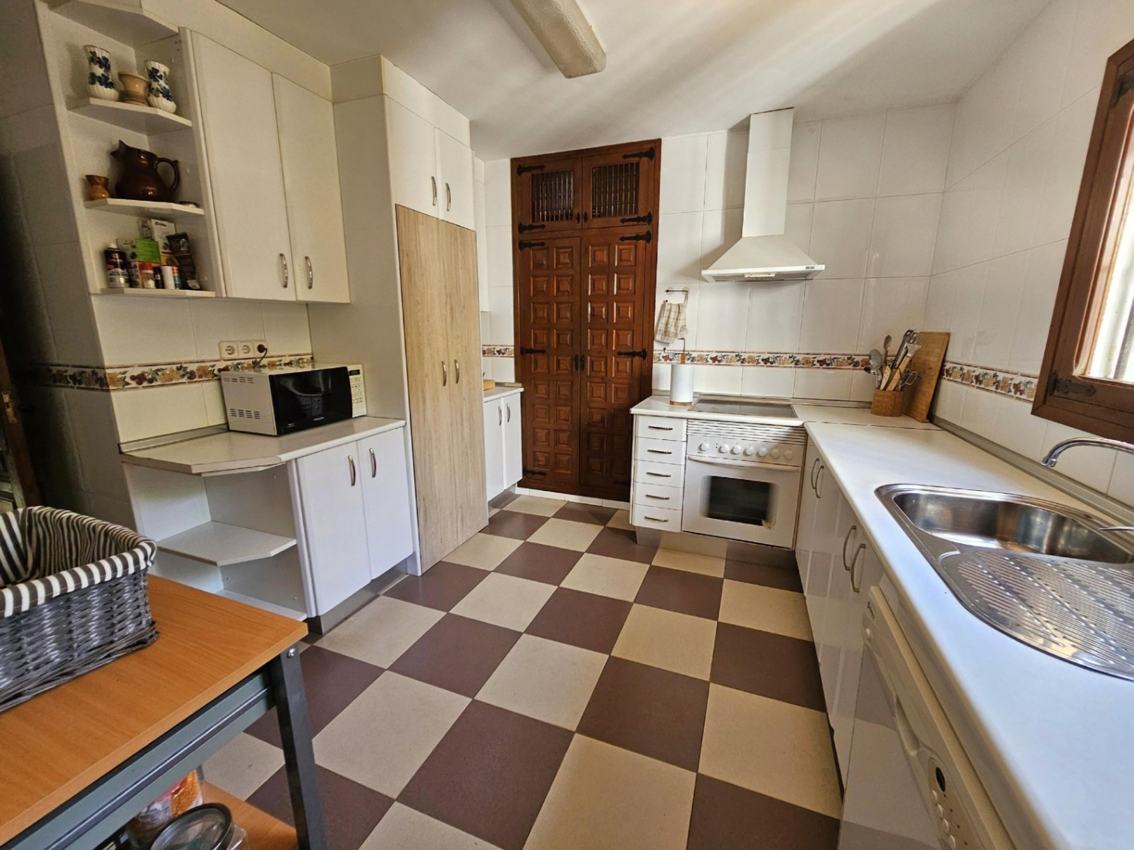 Cocina tradicional con suelo de damero, armarios blancos y una puerta de madera tallada. Incluye fregadero de acero inoxidable y encimeras blancas.