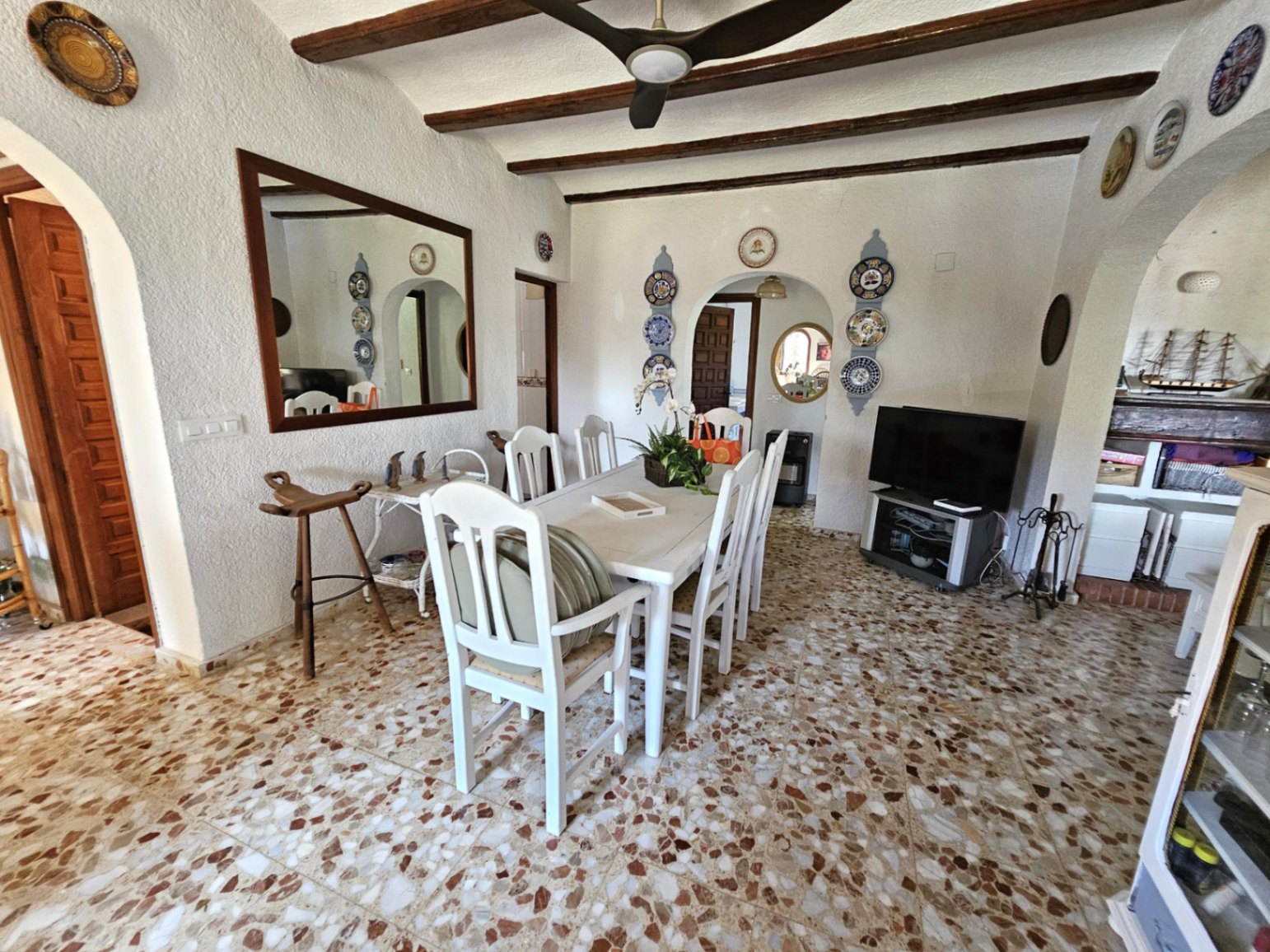 Comedor con vigas de madera a la vista, suelo de terrazo y mobiliario de madera blanca. La decoración incluye platos cerámicos tradicionales y techos con ventilador.