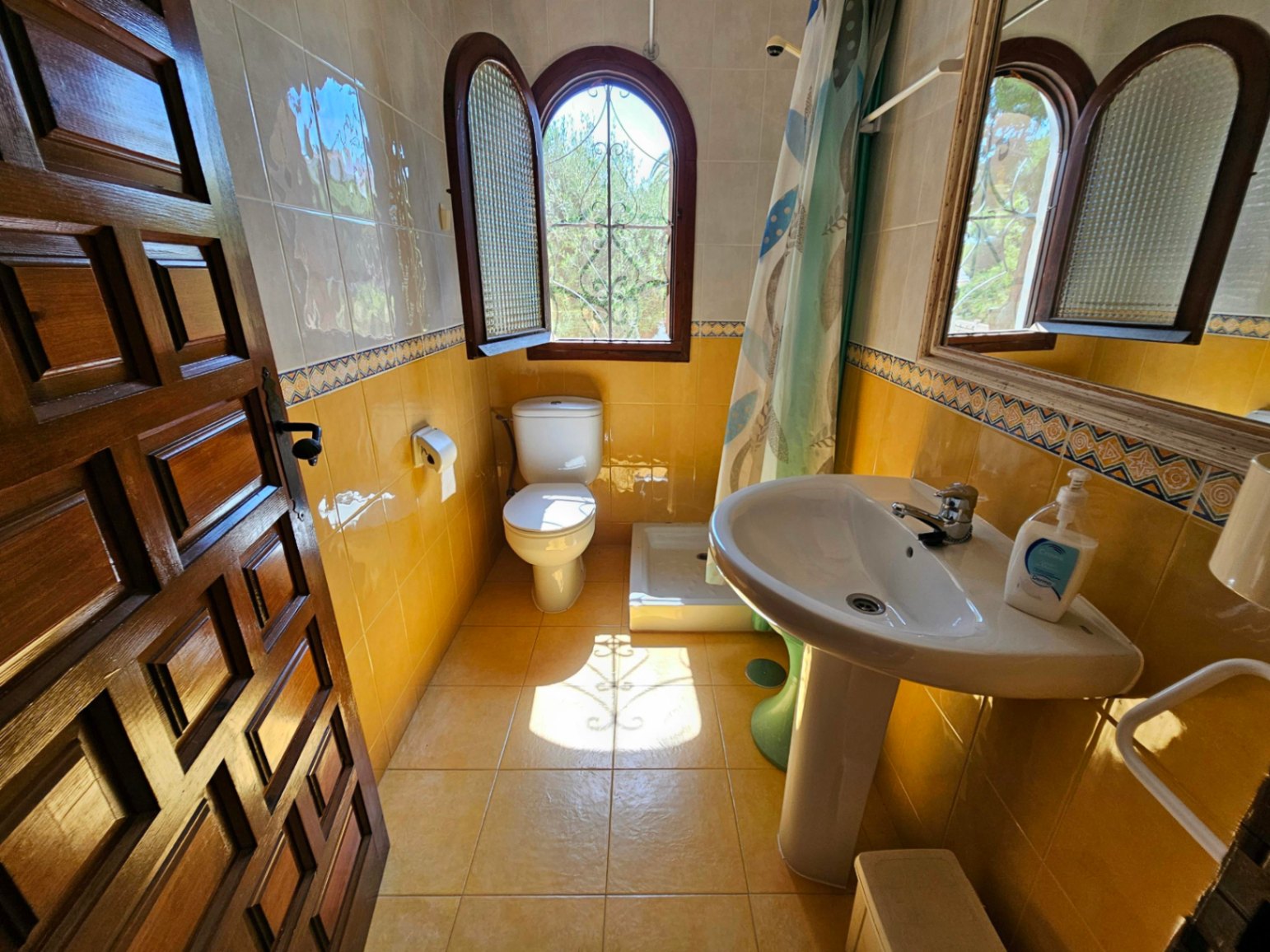 Cuarto de baño con azulejos amarillos y cenefa decorativa, equipado con lavabo de pedestal y ventana de madera en arco con rejería.