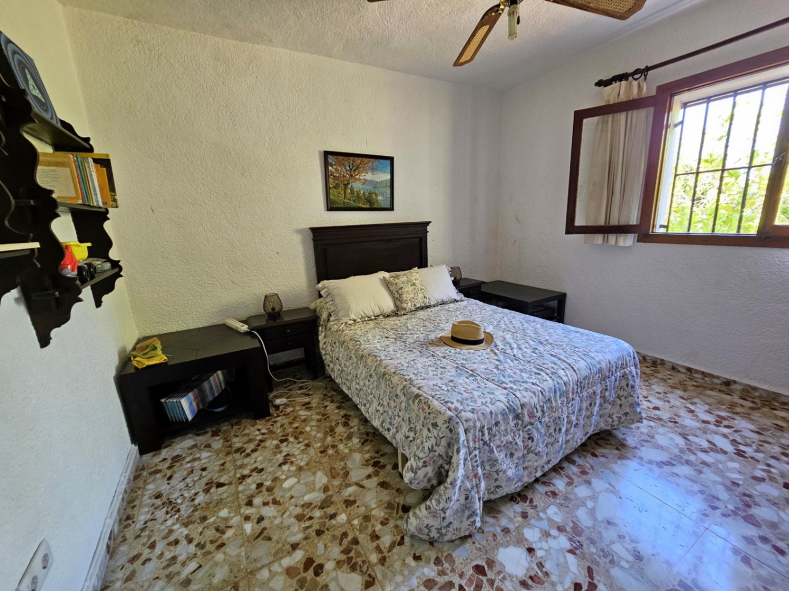 Dormitorio con cama doble, cabecero de madera oscura, suelo de terrazo tradicional y ventana con marco de madera que aporta luz natural.