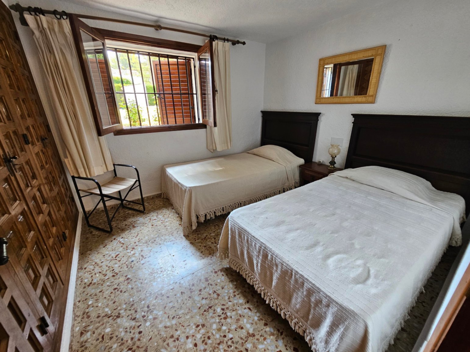 Dormitorio con dos camas individuales, cabeceros de madera oscura y suelo de terrazo. Dispone de armario empotrado de madera y ventana con rejas y contraventanas.