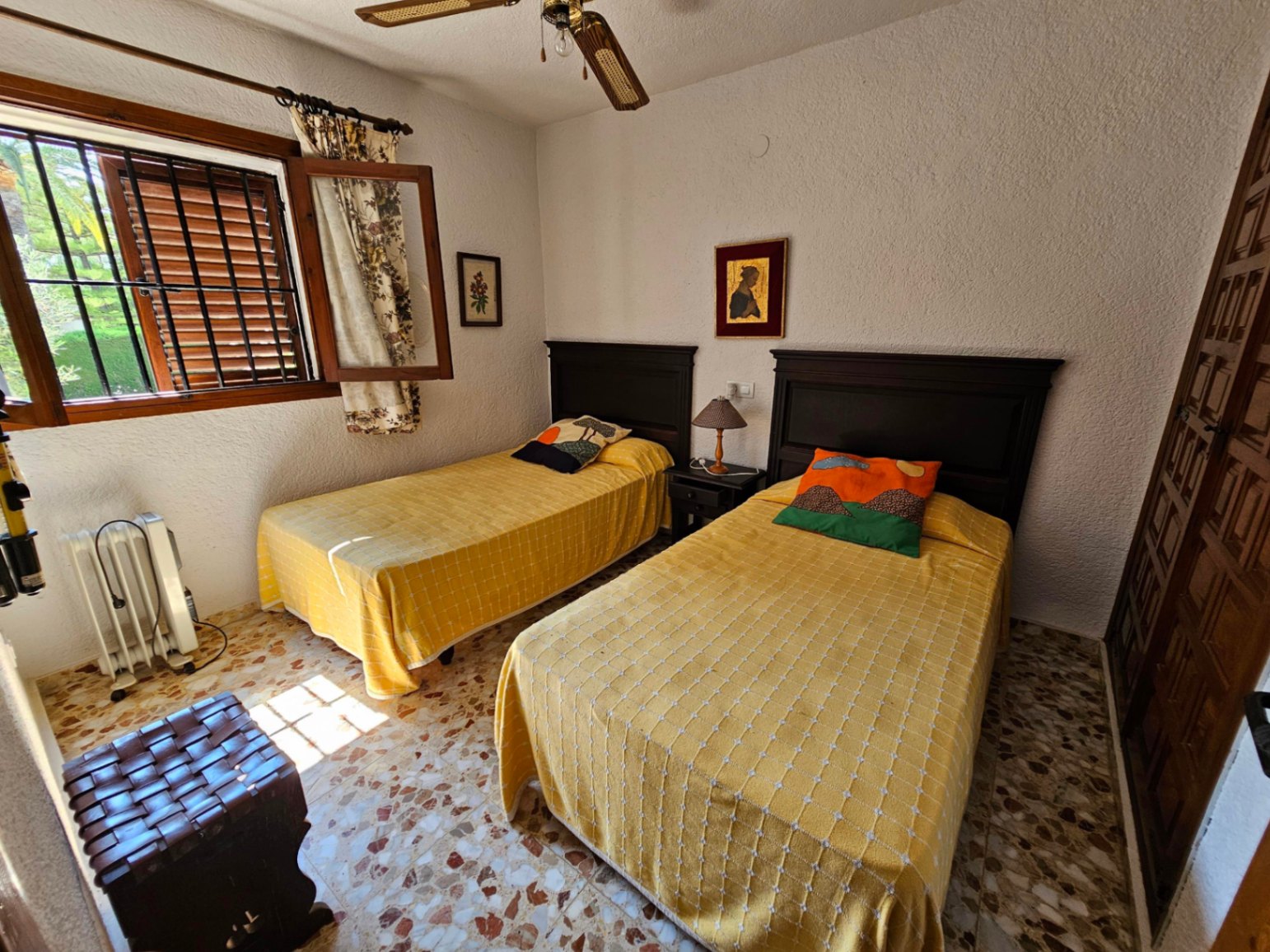 Dormitorio con dos camas individuales, cabeceros de madera oscura y suelo de terrazo. Incluye ventilador de techo y ventana con contraventanas de madera.