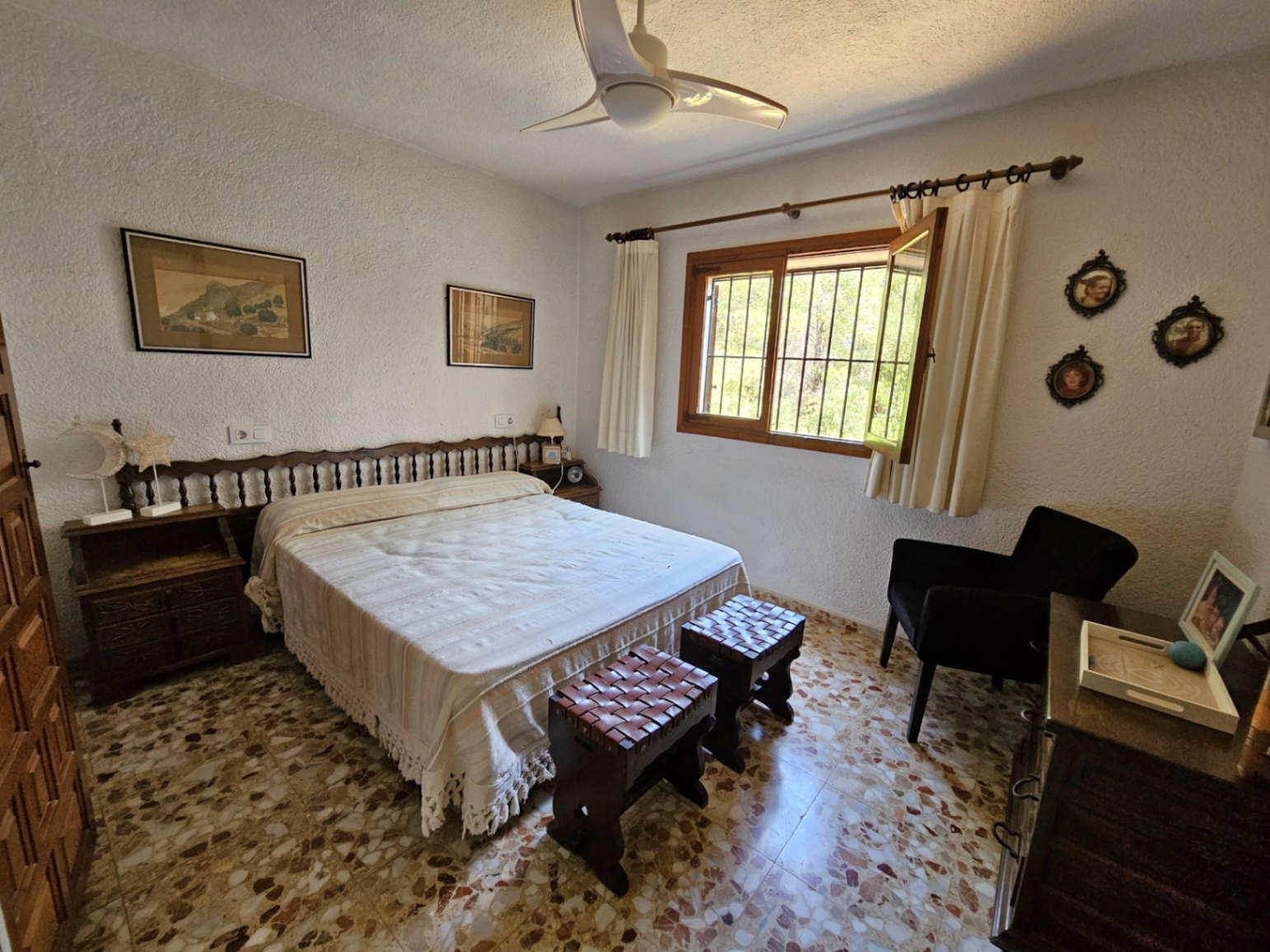 Dormitorio con suelo de terrazo tradicional, cabecero de madera torneada y taburetes de cuero trenzado. Incluye ventilador de techo y ventana de madera con luz natural.