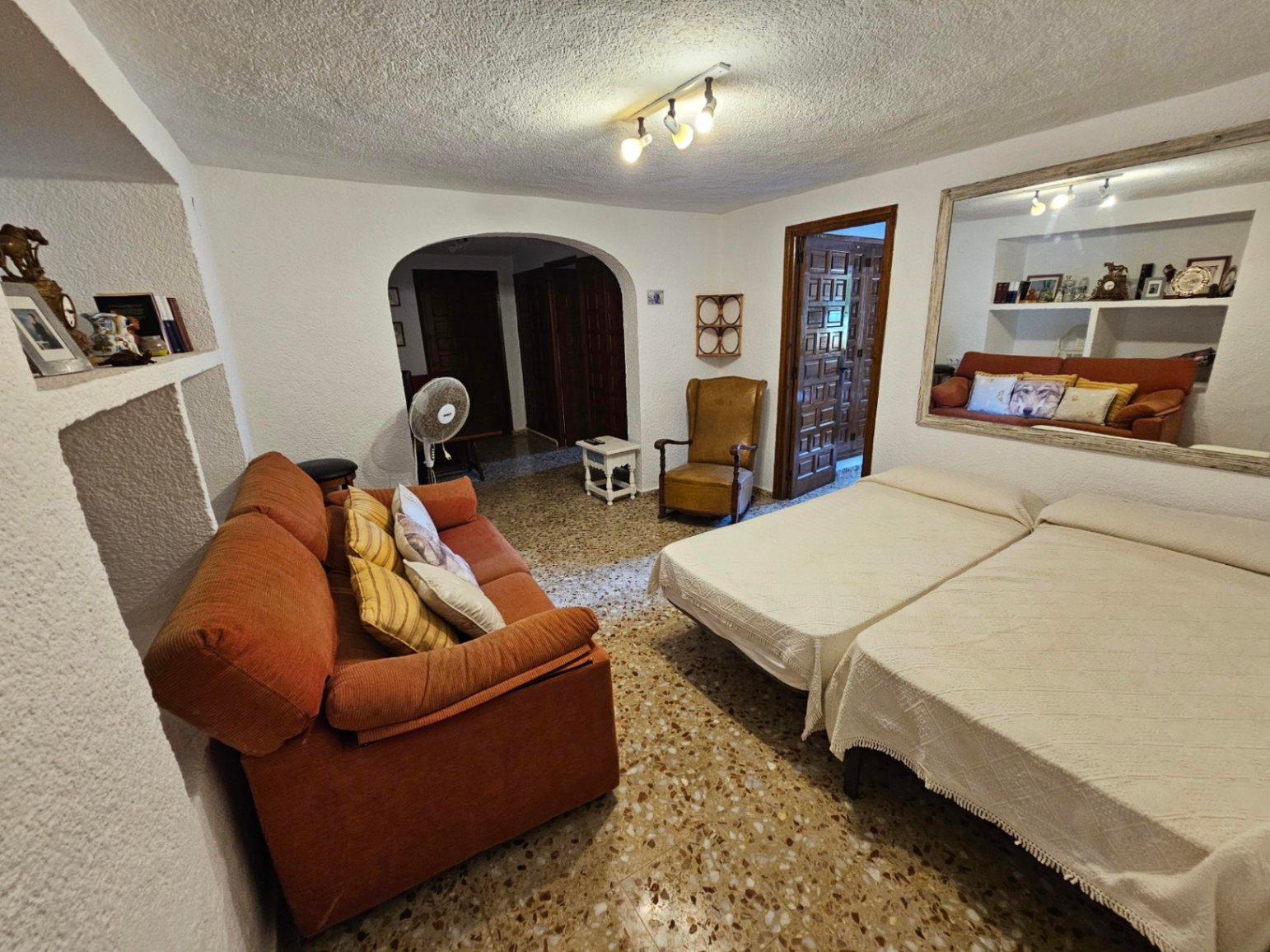 Espacio multifuncional con dos camas individuales, sofá naranja y gran espejo. Destaca el suelo de terrazo, paredes texturizadas y un arco de paso al pasillo.