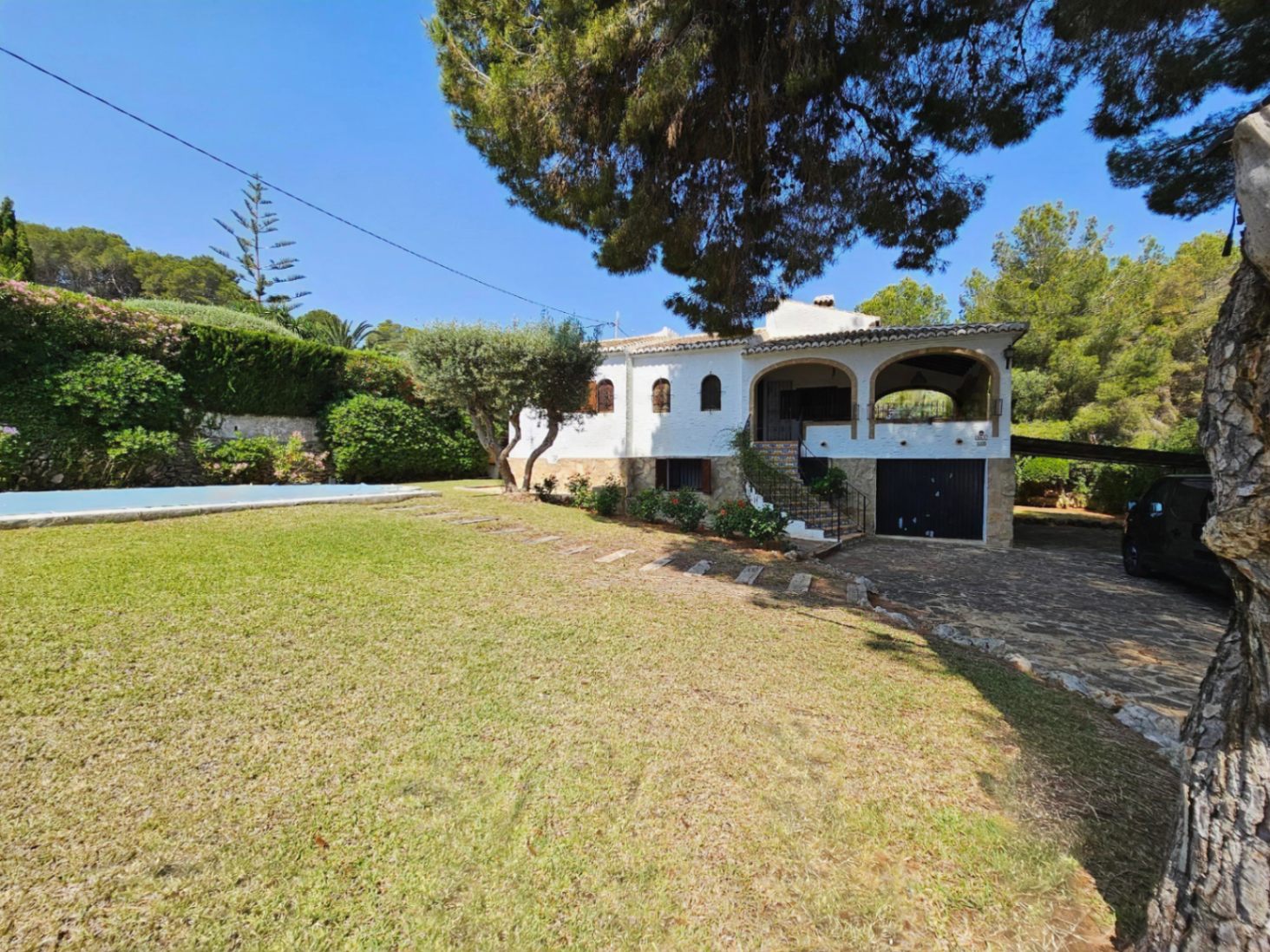 Exterior de una villa mediterránea blanca con porche de arcos, entrada empedrada y jardín con césped junto a la piscina y pinos maduros.