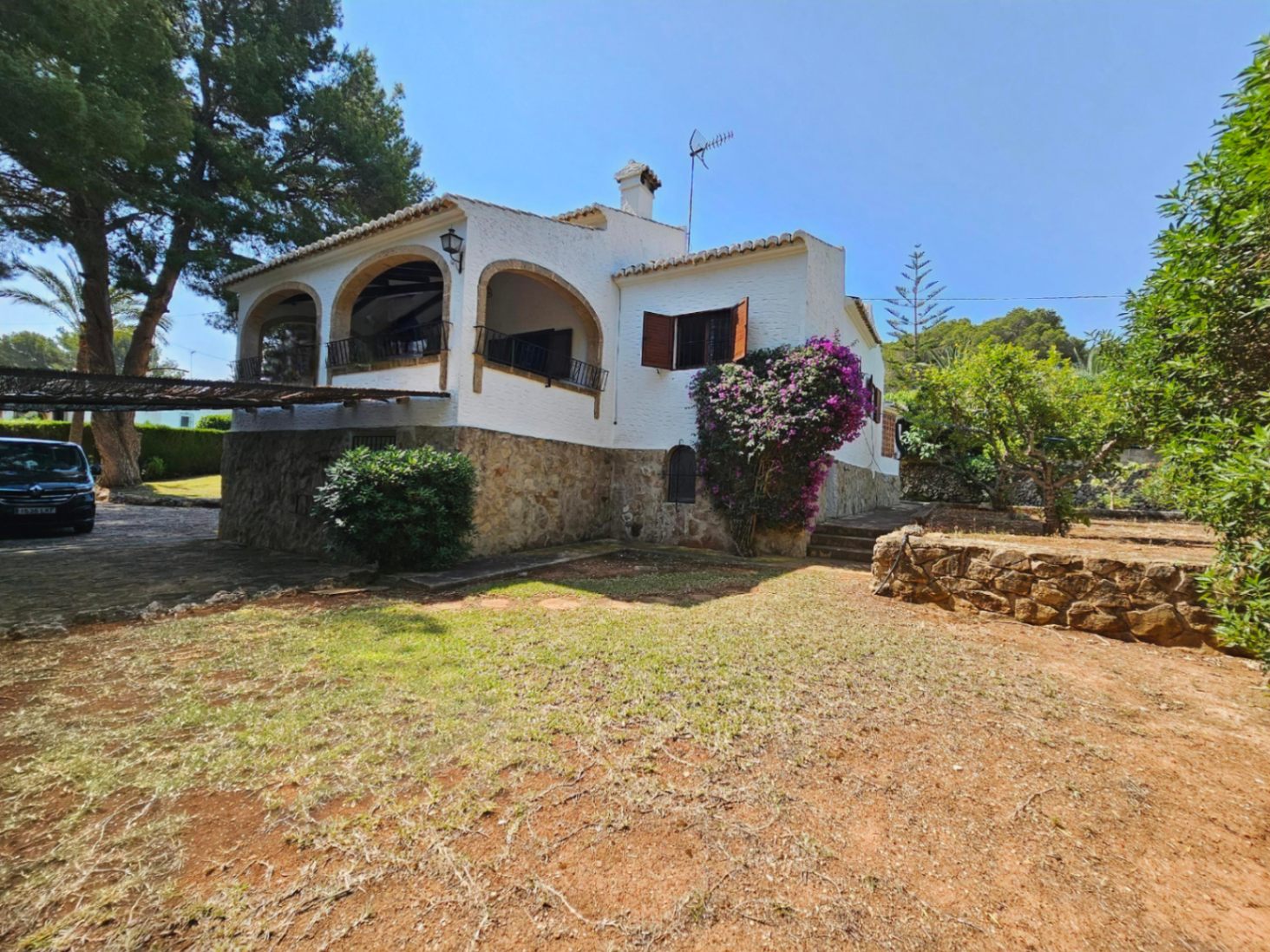 Exterior de villa mediterránea con zócalo de piedra natural, fachada blanca y terraza de arcos. El jardín cuenta con vegetación madura y buganvillas.