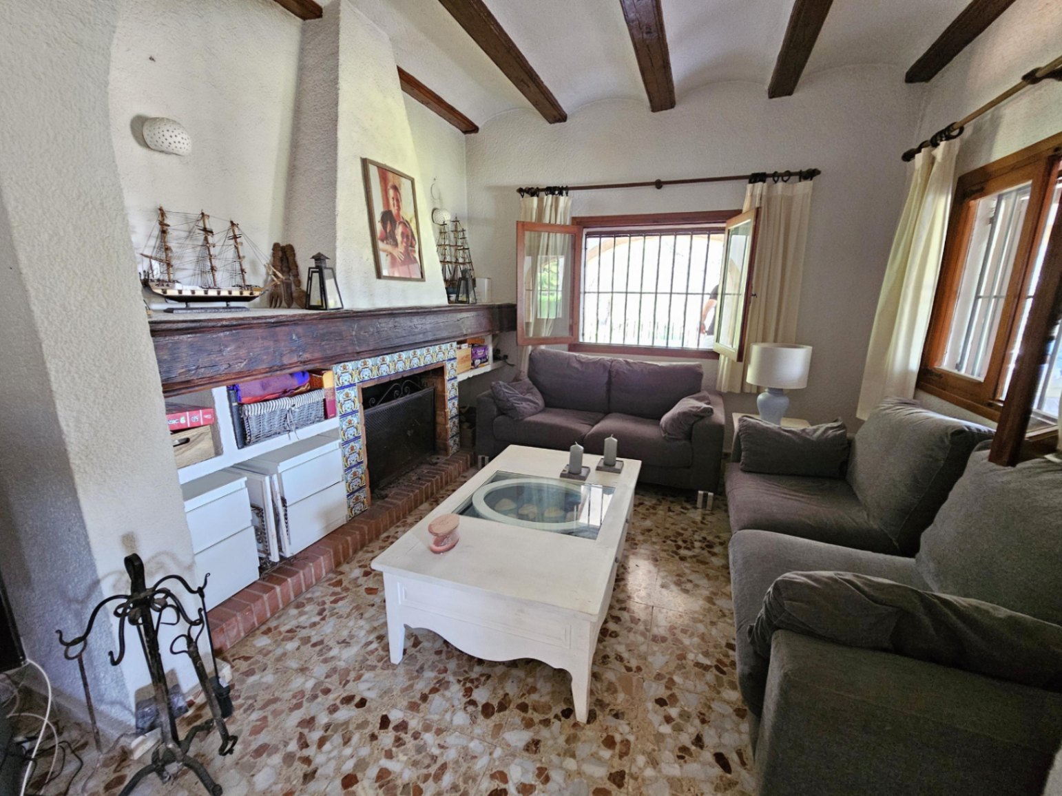 Salón rústico con chimenea de azulejos, viga de madera maciza y techos con vigas vistas. Incluye suelos de terrazo tradicional y mobiliario en tonos neutros.