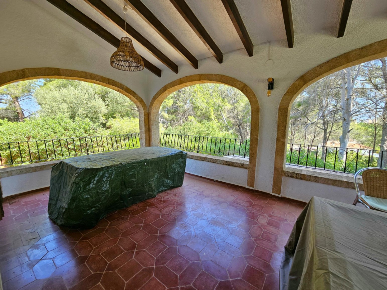 Terraza cubierta con arcos de piedra natural, vigas de madera oscura y suelo de baldosas hexagonales de barro con vistas al entorno arbolado.