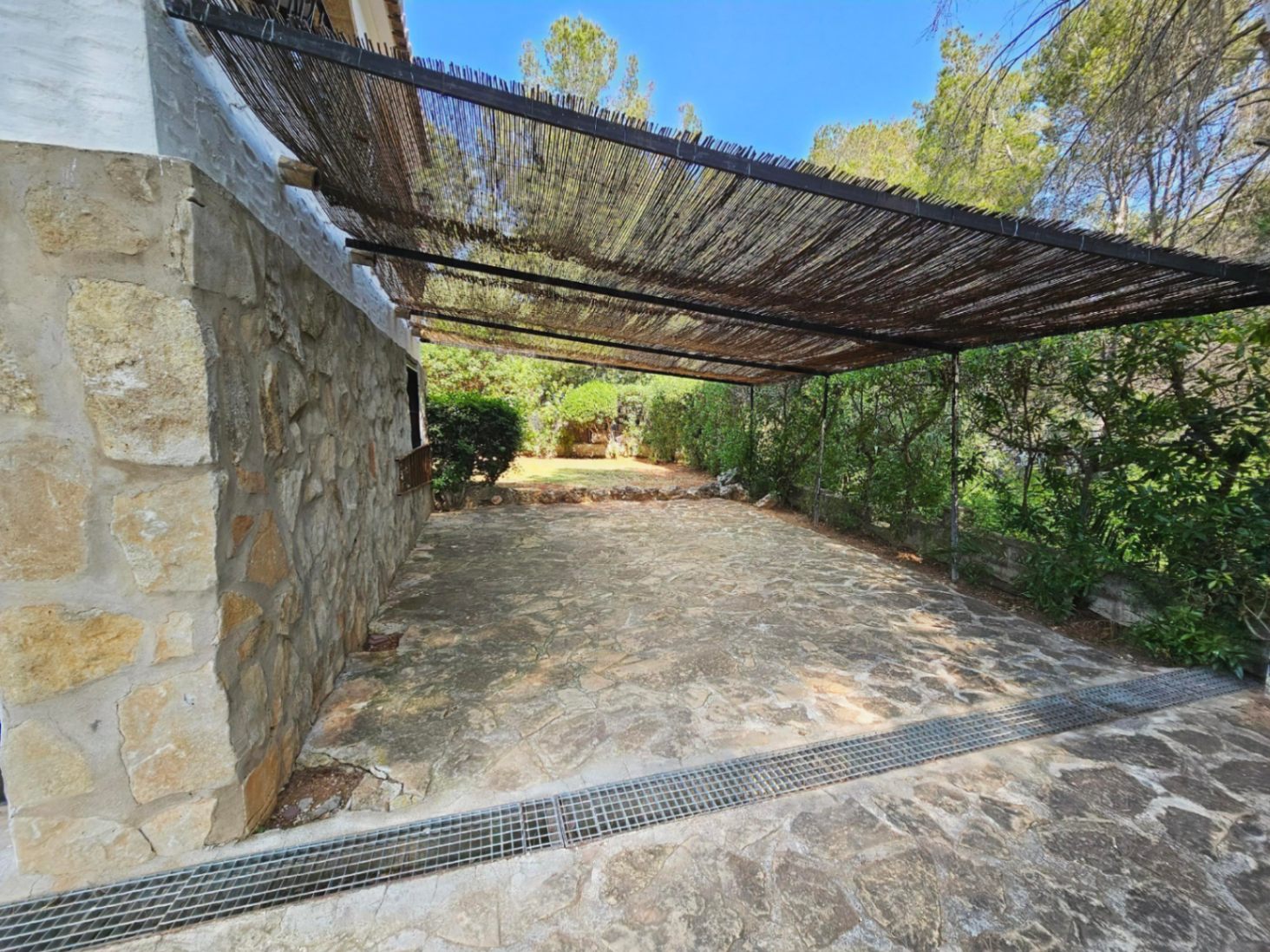 Terraza exterior con suelo de piedra natural y pérgola de cañizo, integrada junto a una fachada de mampostería y rodeada de vegetación mediterránea.