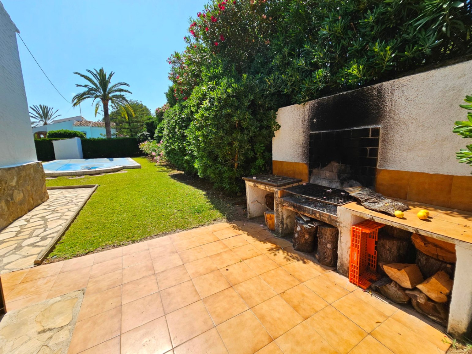 Zona de barbacoa exterior con azulejos de terracota y leñero, con vistas a un jardín de césped cuidado, piscina cubierta y palmeras.