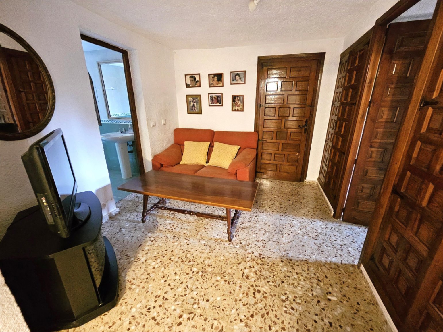Zona de estar con suelo de terrazo tradicional, mesa de centro de madera torneada y puertas castellanas. Incluye un sofá naranja y acceso directo a un baño.