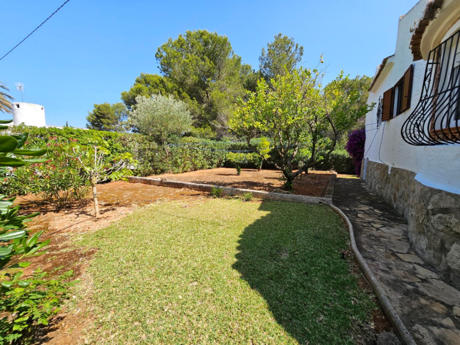 Zona de jardín con césped, árboles frutales y un camino empedrado junto a una casa blanca con zócalo de piedra y contraventanas de madera.