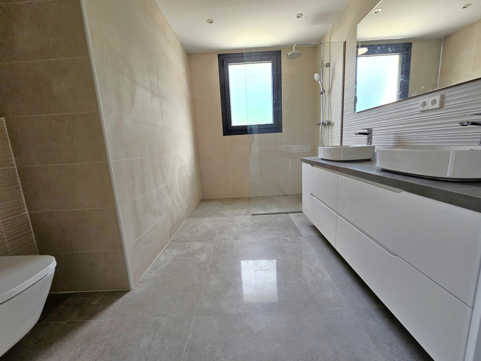 Cuarto de baño moderno con azulejos color crema, tocador blanco con doble lavabo y ducha con mampara de cristal.