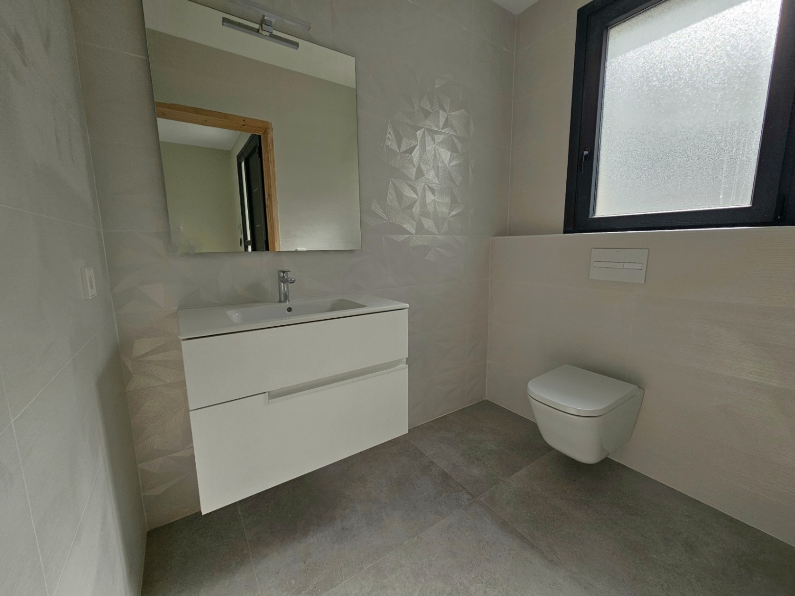Detalle de baño moderno con lavabo suspendido blanco, inodoro y azulejos geométricos.