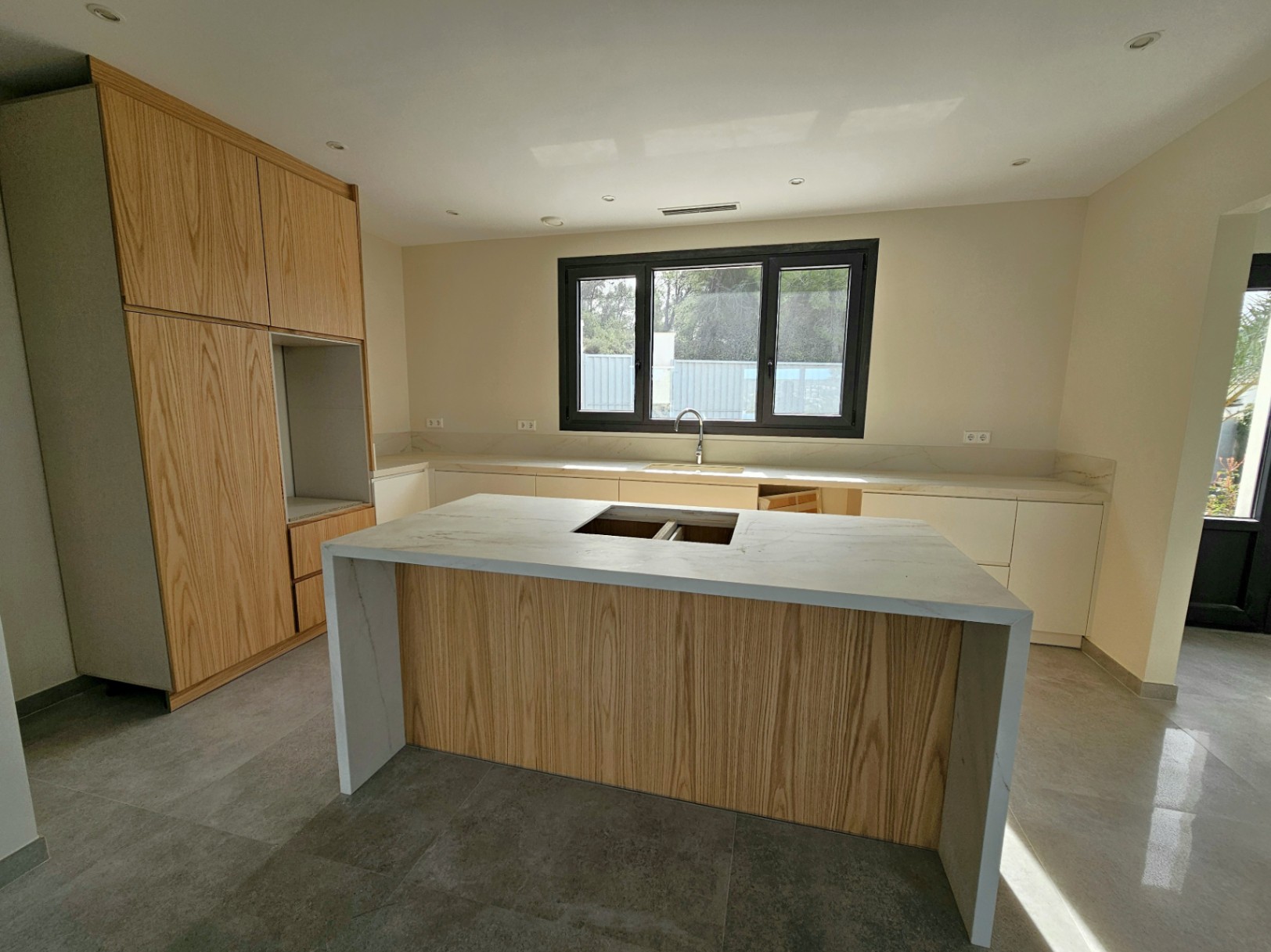 Detalle de la cocina con isla central de madera clara y encimera de piedra veteada, bajo luz natural.