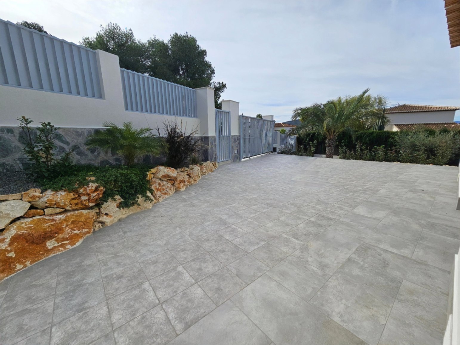 Entrada pavimentada con puerta de seguridad gris y jardín de rocas con plantas mediterráneas.