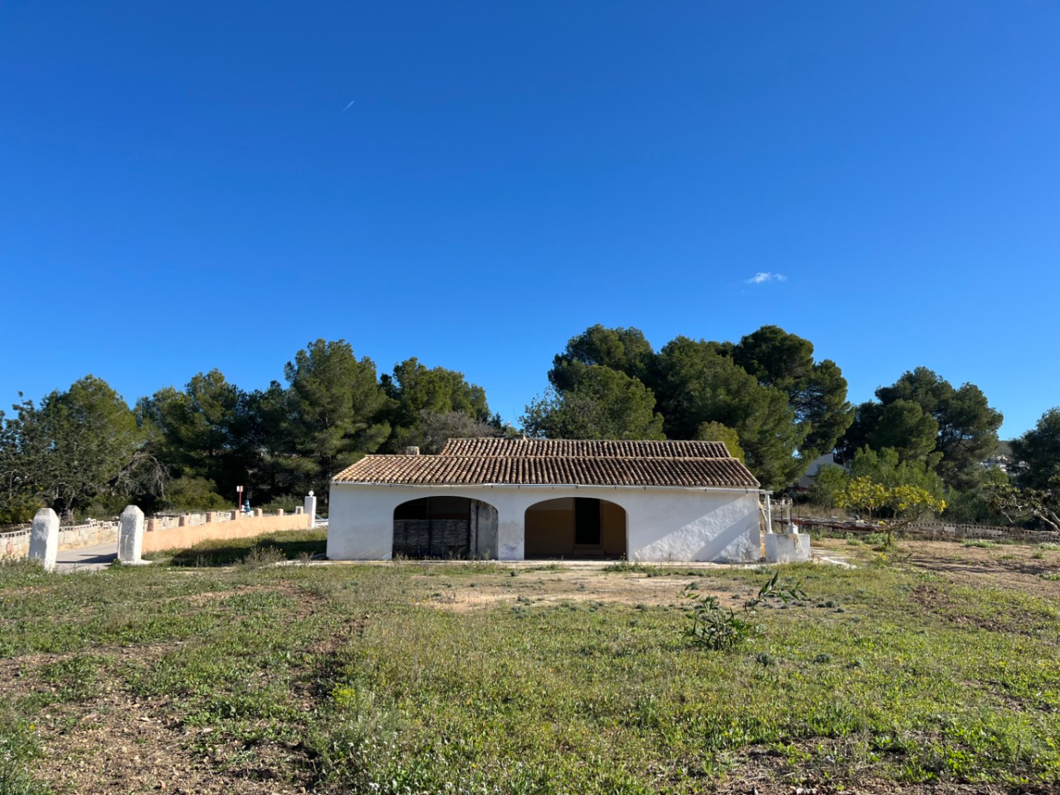 Fachada exterior de una finca tradicional blanca con porche de arcos y tejado de teja árabe, situada en una amplia parcela natural rodeada de pinos.