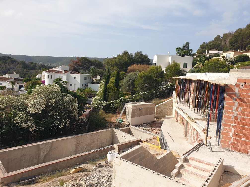 Obra de una villa con cimientos de hormigón, muros de ladrillo con puntales metálicos y excavación para piscina frente a un valle mediterráneo.
