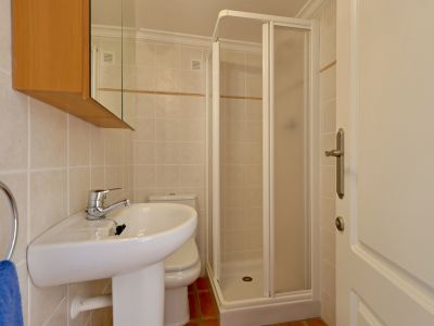 Baño compacto con lavabo de pedestal, inodoro y cabina de ducha angular. Presenta azulejos claros con cenefa decorativa y suelo de terracota.