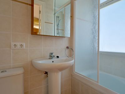 Baño moderno con lavabo de pedestal blanco, inodoro y paredes de azulejos claros. Un espejo-armario cuelga sobre el lavabo, junto a una bañera con mampara de cristal esmerilado.
