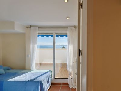 Dormitorio luminoso con cama azul y acceso directo a una terraza con vistas panorámicas al mar Mediterráneo. Suelo de terracota y cortinas blancas.