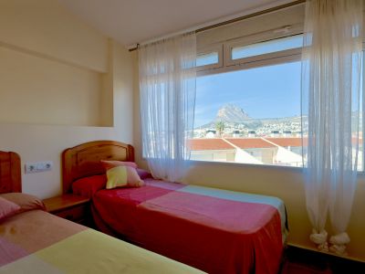 Dormitorio luminoso con dos camas individuales de cabecero de madera, ofreciendo una amplia ventana con vistas a la montaña del Montgó y tejados.