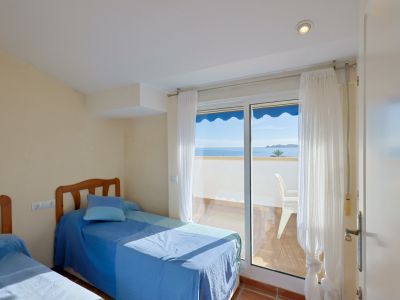 Dormitorio luminoso con dos camas individuales y acceso directo a un balcón privado que ofrece vistas panorámicas al mar y a la costa. Suelo de baldosas de terracota y abundante luz natural.