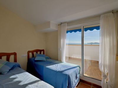 Dormitorio luminoso con dos camas individuales y cabeceros de madera. Acceso a un balcón con suelo de terracota, vistas al mar y un toldo azul.