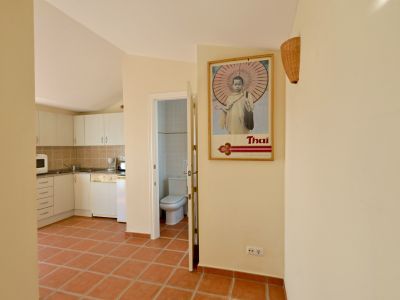 Pasillo con suelo de baldosas de terracota que conecta la cocina abierta con muebles blancos y un baño visible a través de una puerta entreabierta. Un póster 'Thai' decora la pared.