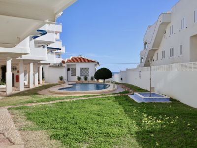 Piscina comunitaria de forma orgánica con mosaico azul, rodeada por césped y edificios blancos de estilo mediterráneo. Al fondo se vislumbra el mar bajo un cielo despejado.