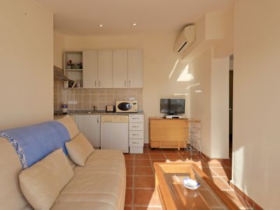 Salón-comedor diáfano con cocina integrada, suelo de terracota y armarios blancos. La zona de estar incluye un sofá claro y mesa de centro de cristal.