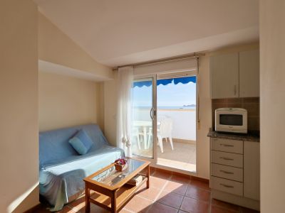 Salón-comedor funcional con sofá y mesa de centro, que integra una cocina compacta. Acceso directo a una terraza con mobiliario exterior y vistas panorámicas al mar.