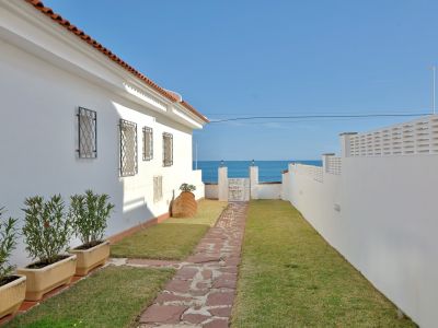 Sendero de piedra que atraviesa un césped cuidado hacia una verja blanca, revelando vistas directas al mar bajo un cielo azul claro. Un edificio blanco con tejado de teja y ventanas enrejadas a la izquierda.