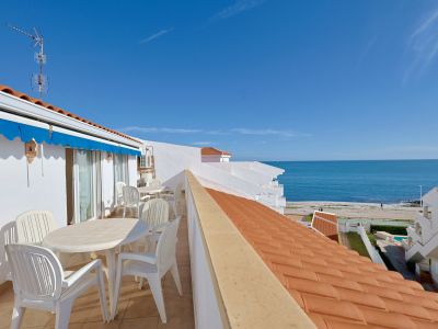Terraza exterior con mobiliario blanco, toldo azul y vistas directas al mar Mediterráneo y la playa, bajo un cielo despejado.