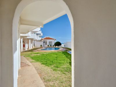 Vista a través de un arco blanco que enmarca una piscina comunitaria con césped, un edificio de tejado de teja y el mar Mediterráneo al fondo bajo un cielo azul.
